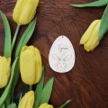 Drewniana zawieszka w kształcie jaja z grawerem imienia Tymon i zajączkiem, otoczona żółtymi tulipanami na drewnianym tle.