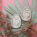 Drewniane zawieszki wielkanocne w kształcie jajek, z napisem Happy Easter i Wesołego Alleluja, z motywem zajączka, na tle dekoracji z liści i kwiatów.