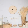 Okrągły drewniany zegar na ścianę w jasnym kolorze. Obok dekoracje: wazon z suszonymi kwiatami, szklane pojemniki, metalowy stojak na biżuterię.