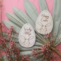 Dwie drewniane zawieszki Happy Easter w kształcie jajka z grawerunkiem królika i napisem Happy Easter, na tle dekoracji.