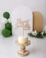 Topper na tort Chrzest Święty ze sklejki brzozowej wbity w kremowy tort, stojący na drewnianej paterze.